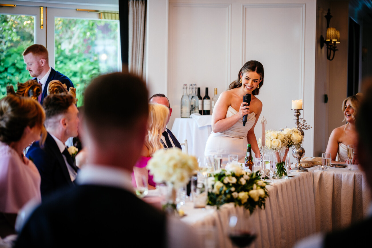 Tara and Darren’s Unforgettable Wedding at Bellingham Castle 150 A bridesmaid gives a speech into a microphone during the wedding reception at Bellingham Castle, surrounded by floral centrepieces and candles.