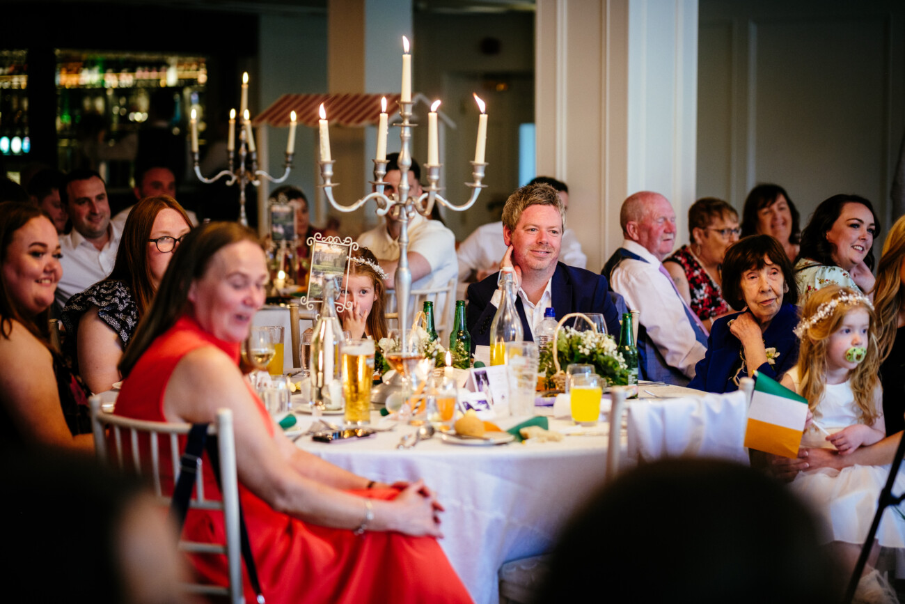 Tara and Darren’s Unforgettable Wedding at Bellingham Castle 151 A group of people sit around a decorated table at an indoor event, smiling and watching something off camera; a child holds an Irish flag, capturing the joyful spirit of a wedding at Bellingham Castle.
