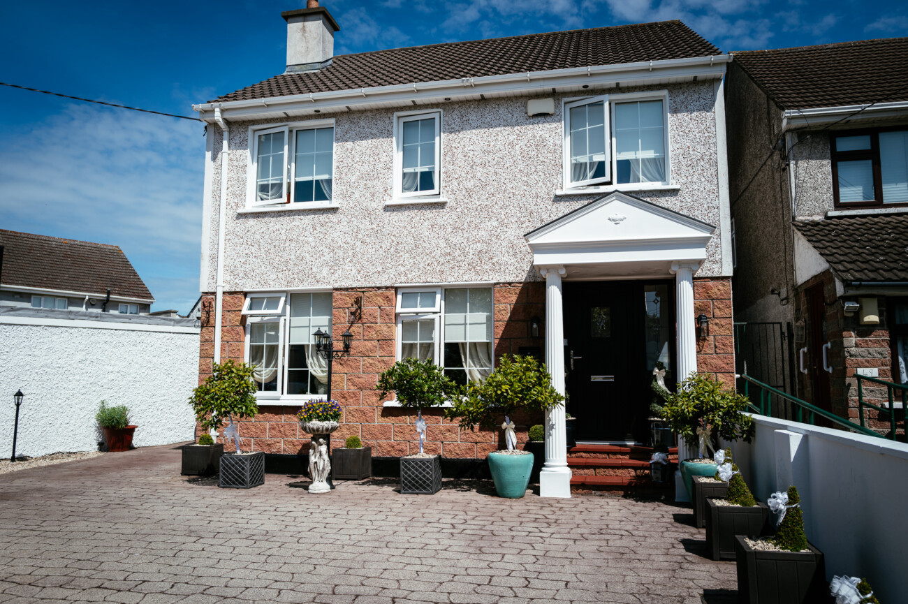 Tara and Darren’s Unforgettable Wedding at Bellingham Castle 1 Two-storey house with white walls, brown roof, and front porch. Several potted plants and statues line the entrance on a paved drive, evoking the elegance of a wedding at Bellingham Castle.