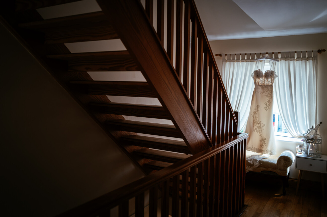 Tara and Darren’s Unforgettable Wedding at Bellingham Castle 2 A lace wedding dress hangs on a hanger in front of a window with sheer curtains, next to a small sofa and a side table, seen through a wooden staircase—capturing the elegant anticipation of a wedding at Bellingham Castle.