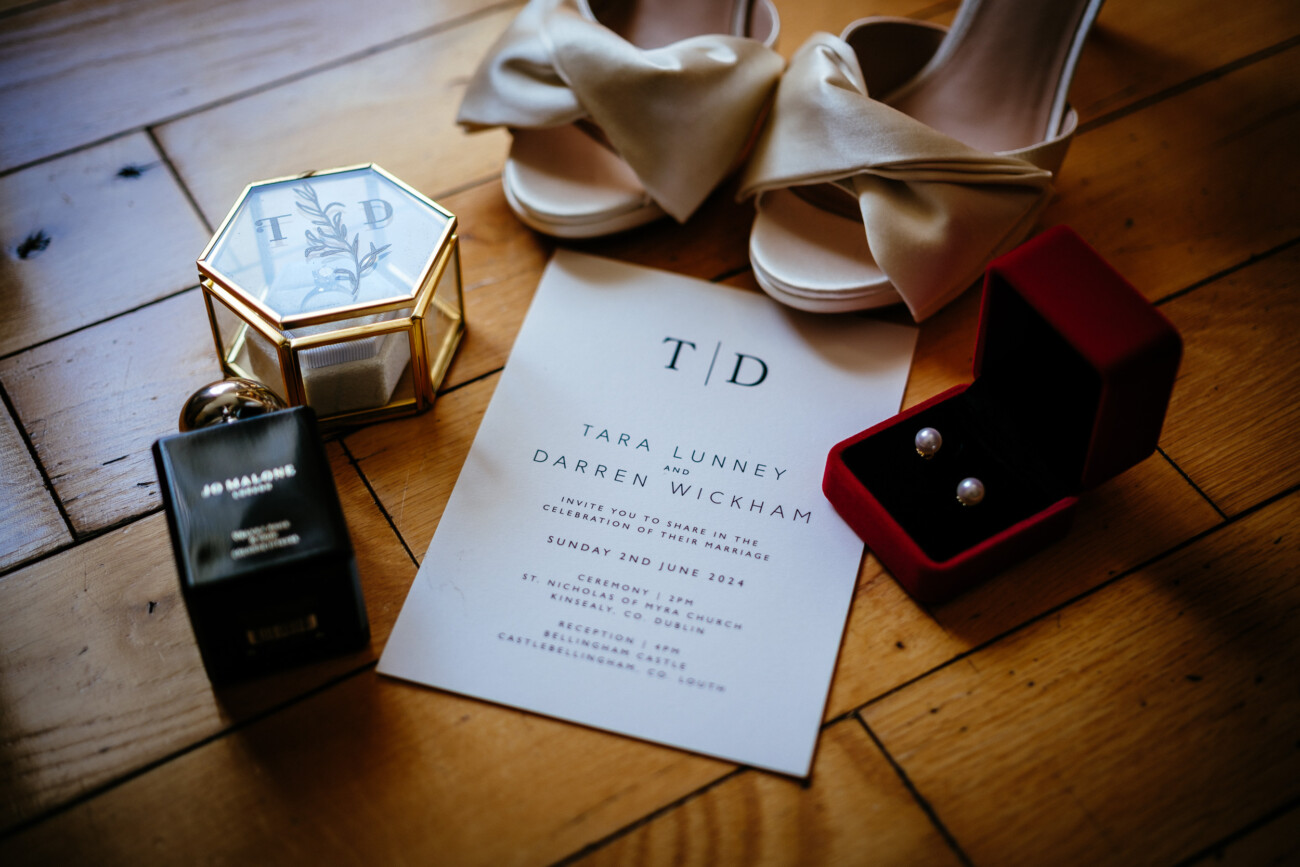 Tara and Darren’s Unforgettable Wedding at Bellingham Castle 3 Tara and Darren's wedding invitation with their initials on a keepsake box, pearl earrings, perfume, and the bride's shoes, detailing their ceremony at St. Nicholas of Myra Church and reception at Bellingham Castle.