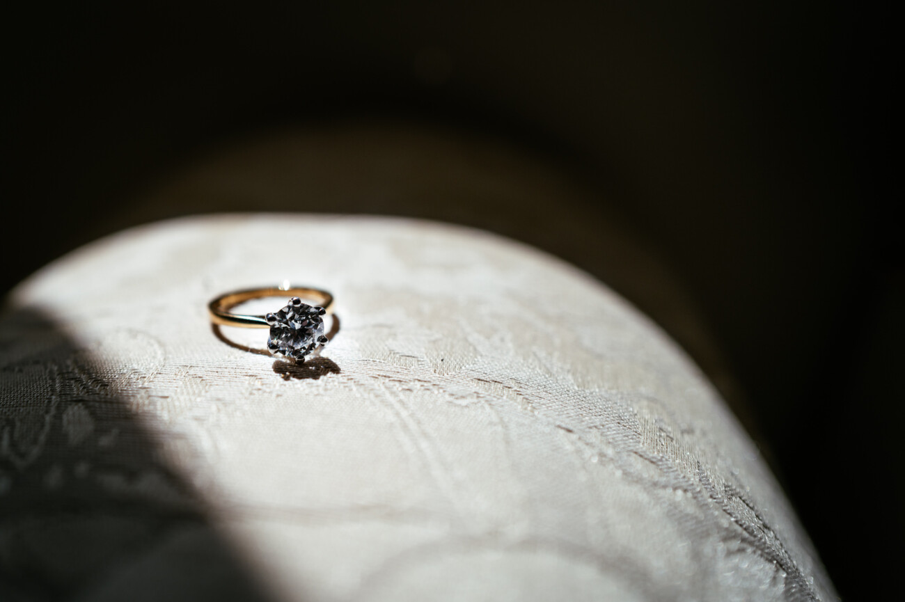 Tara and Darren’s Unforgettable Wedding at Bellingham Castle 4 A gold ring with a single large diamond is placed on a textured light fabric, illuminated by a beam of sunlight against a dark background—capturing the elegance and romance of a wedding at Bellingham Castle.