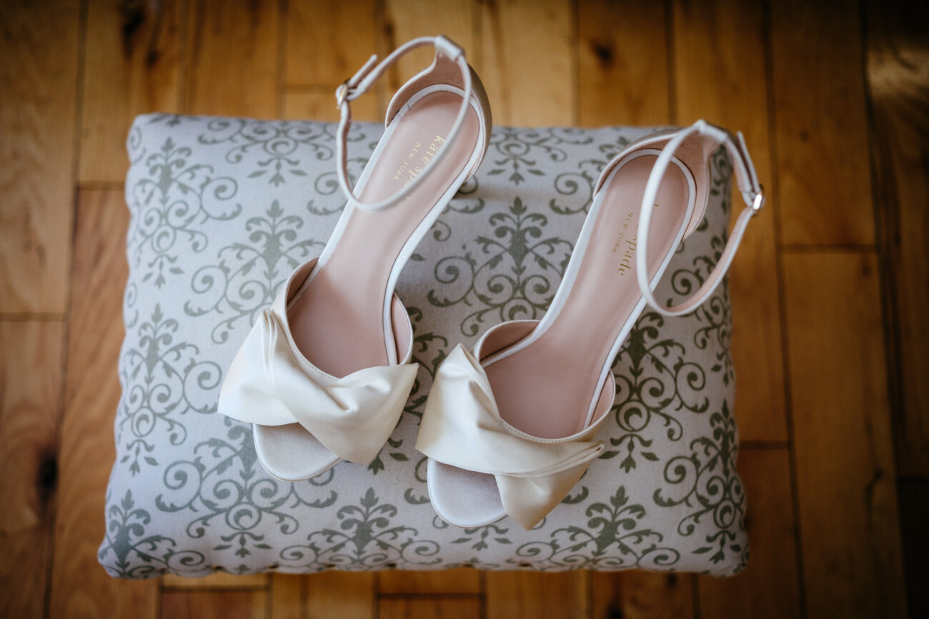 Tara and Darren’s Unforgettable Wedding at Bellingham Castle 6 A pair of white open-toe heeled sandals with bows on the front, placed on a patterned cushion on a wooden floor—perfect for a romantic wedding at Bellingham Castle.