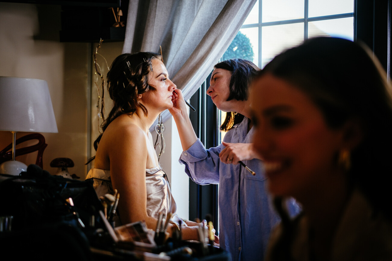 Tara and Darren’s Unforgettable Wedding at Bellingham Castle 8 A woman sits as a make-up artist applies make-up to her face near a window, preparing for her wedding at Bellingham Castle, whilst another woman is blurred in the foreground.