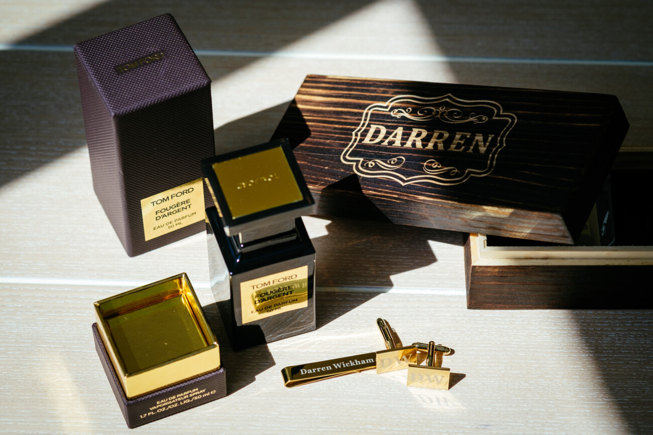 Tara and Darren’s Unforgettable Wedding at Bellingham Castle 10 A Tom Ford perfume bottle and box sit next to a gold key, a brass nameplate, and a wooden box engraved with “DARREN” on a sunlit surface—evoking the refined elegance of a wedding at Bellingham Castle.