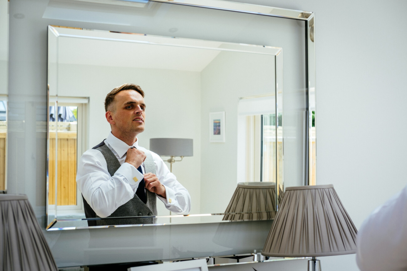 Tara and Darren’s Unforgettable Wedding at Bellingham Castle 13 A man adjusts his tie in front of a large mirror in a bright, modern room with lamps and framed pictures, preparing for a wedding at Bellingham Castle.