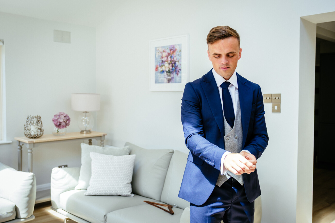 Tara and Darren’s Unforgettable Wedding at Bellingham Castle 15 A man in a blue suit adjusts his cuff while standing in a bright living room with light-coloured furniture and wall art, preparing for a wedding at Bellingham Castle.