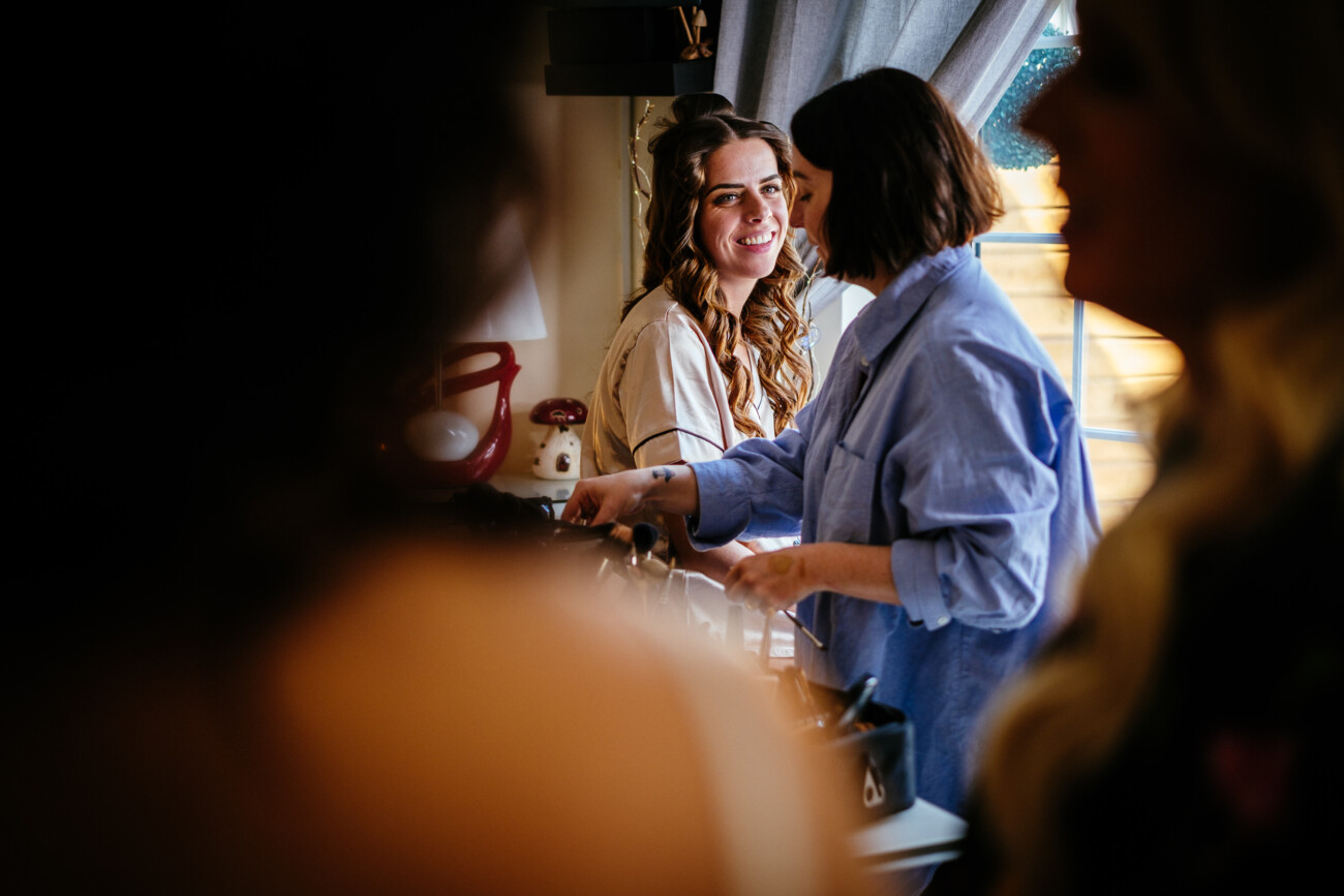Tara and Darren’s Unforgettable Wedding at Bellingham Castle 17 A woman with curled hair smiles whilst another woman, possibly a make-up artist, prepares cosmetics on a table near a window, capturing the joyful preparations for a wedding at Bellingham Castle.