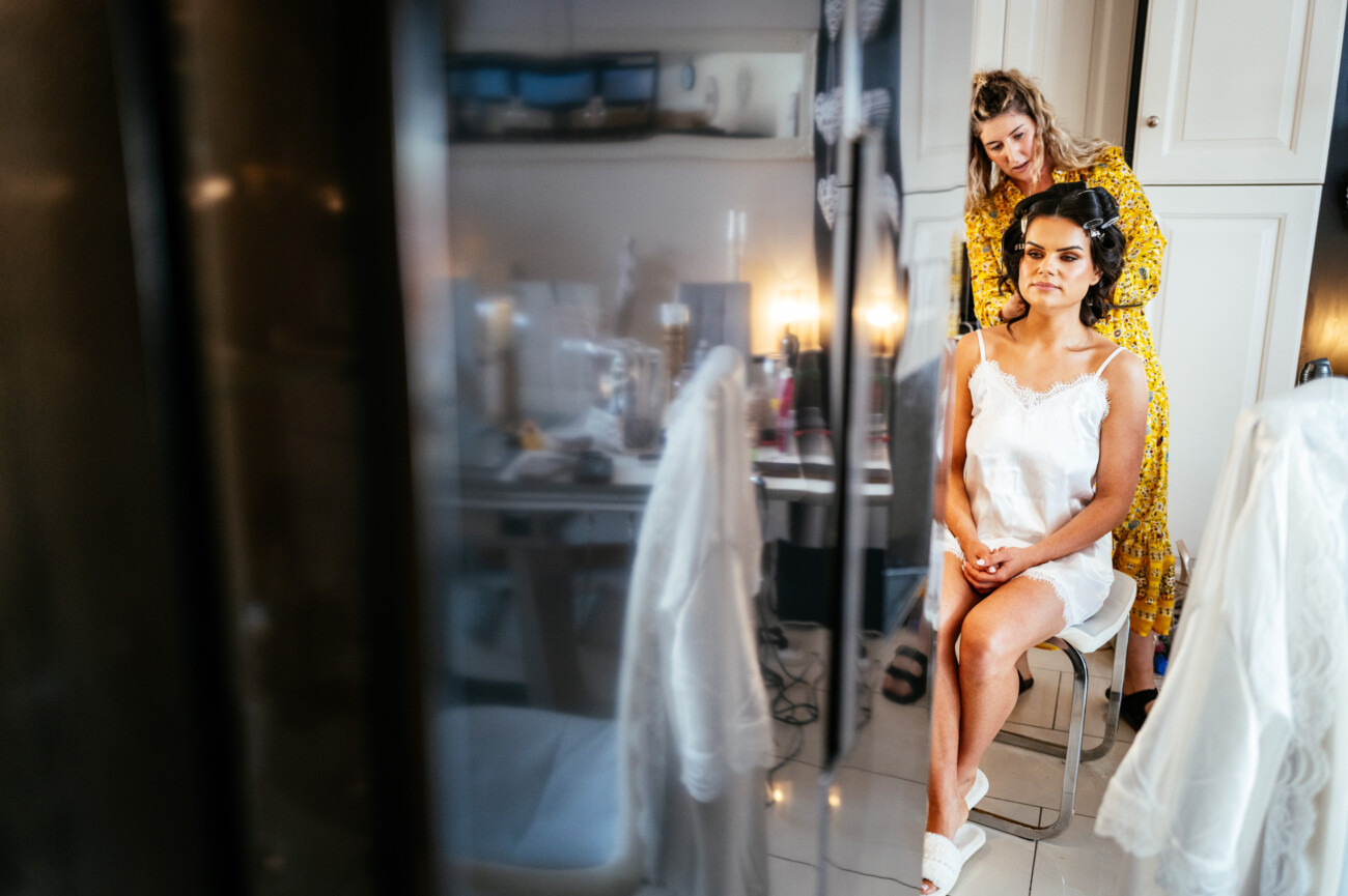 Tara and Darren’s Unforgettable Wedding at Bellingham Castle 19 A woman sits on a chair in a white outfit whilst another woman styles her hair in a bright, modern room with mirrors and cupboards, preparing for her wedding at Bellingham Castle.