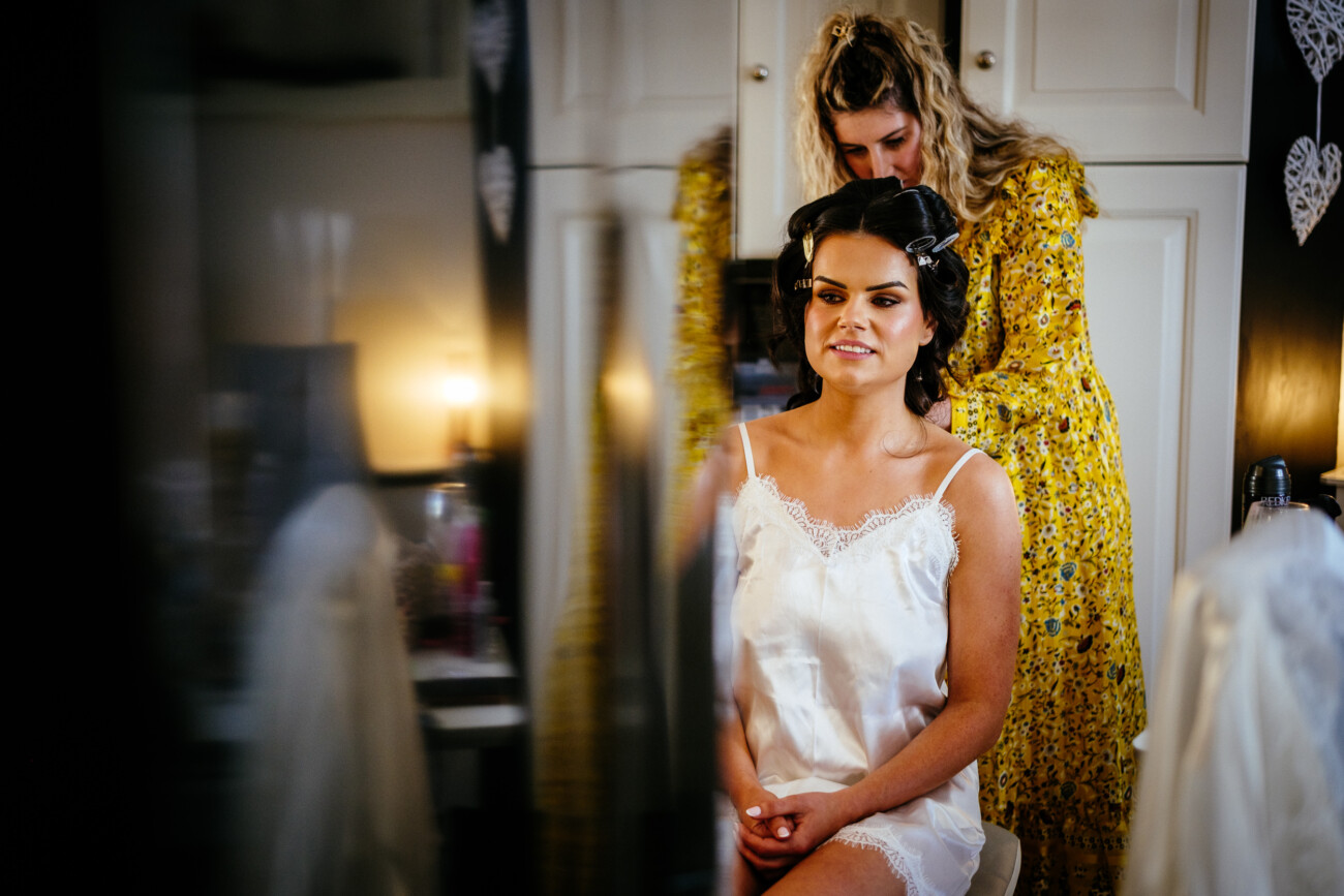 Tara and Darren’s Unforgettable Wedding at Bellingham Castle 20 A woman wearing a white satin petticoat sits while another woman in a yellow dress styles her hair in a well-lit room, preparing for the wedding at Bellingham Castle.