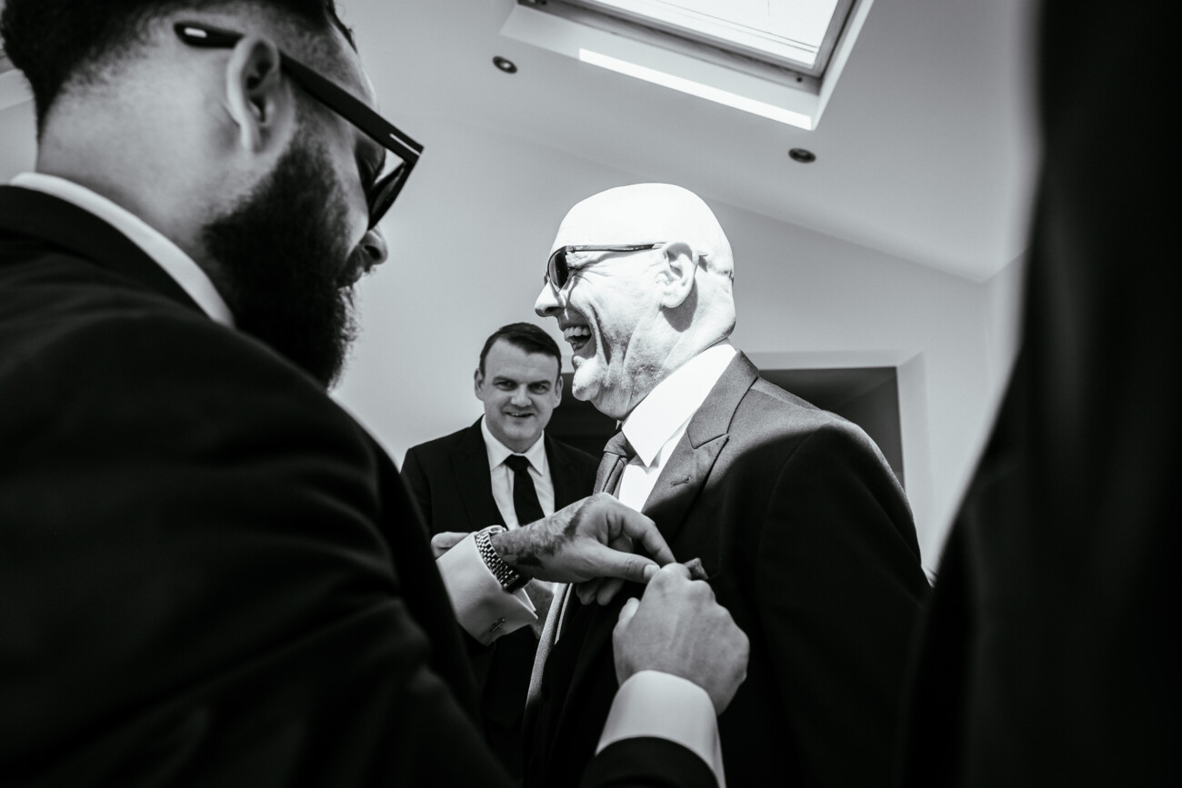 Tara and Darren’s Unforgettable Wedding at Bellingham Castle 23 Three men in suits indoors, one attaching a buttonhole to another’s lapel—all wearing glasses, with one man smiling. This black and white photo beautifully captures a candid moment during a wedding at Bellingham Castle.