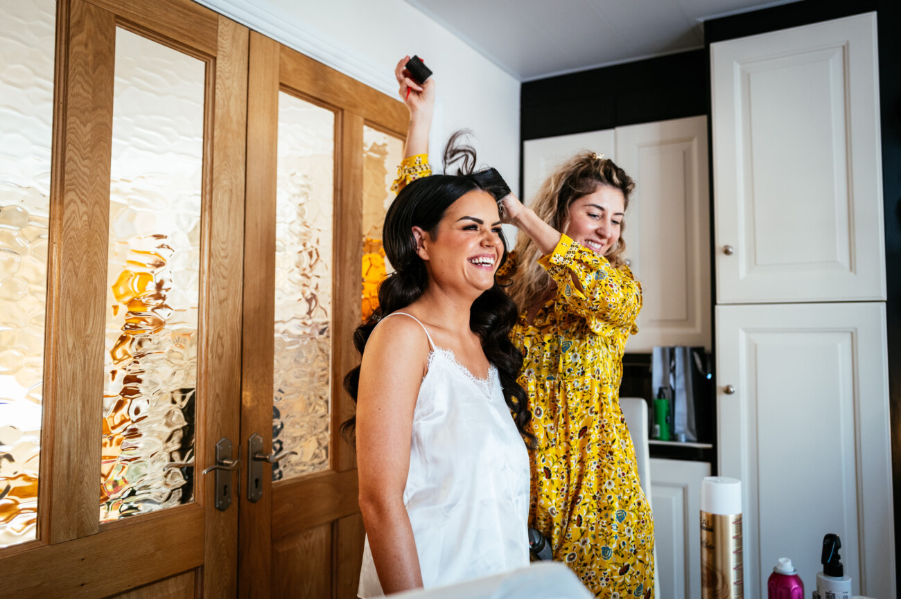 Tara and Darren’s Unforgettable Wedding at Bellingham Castle 24 A woman in a white top smiles as another woman in a yellow floral dress styles her hair in a brightly lit room, preparing for the wedding at Bellingham Castle.