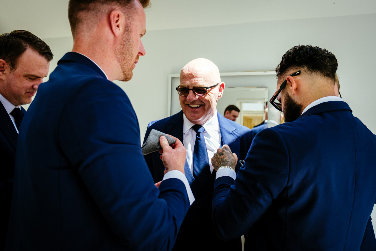 Tara and Darren’s Unforgettable Wedding at Bellingham Castle 25 Four men in suits stand together indoors, adjusting ties and pocket squares, with one man smiling in the centre of the group as they prepare for a wedding at Bellingham Castle.