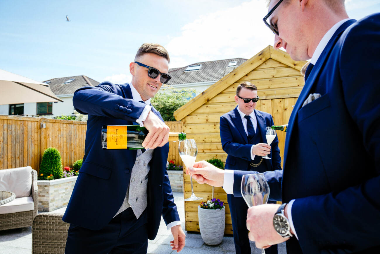 Tara and Darren’s Unforgettable Wedding at Bellingham Castle 26 Three men in suits and sunglasses pour champagne into glasses outdoors near a wooden shed on a sunny day, celebrating a wedding at Bellingham Castle.