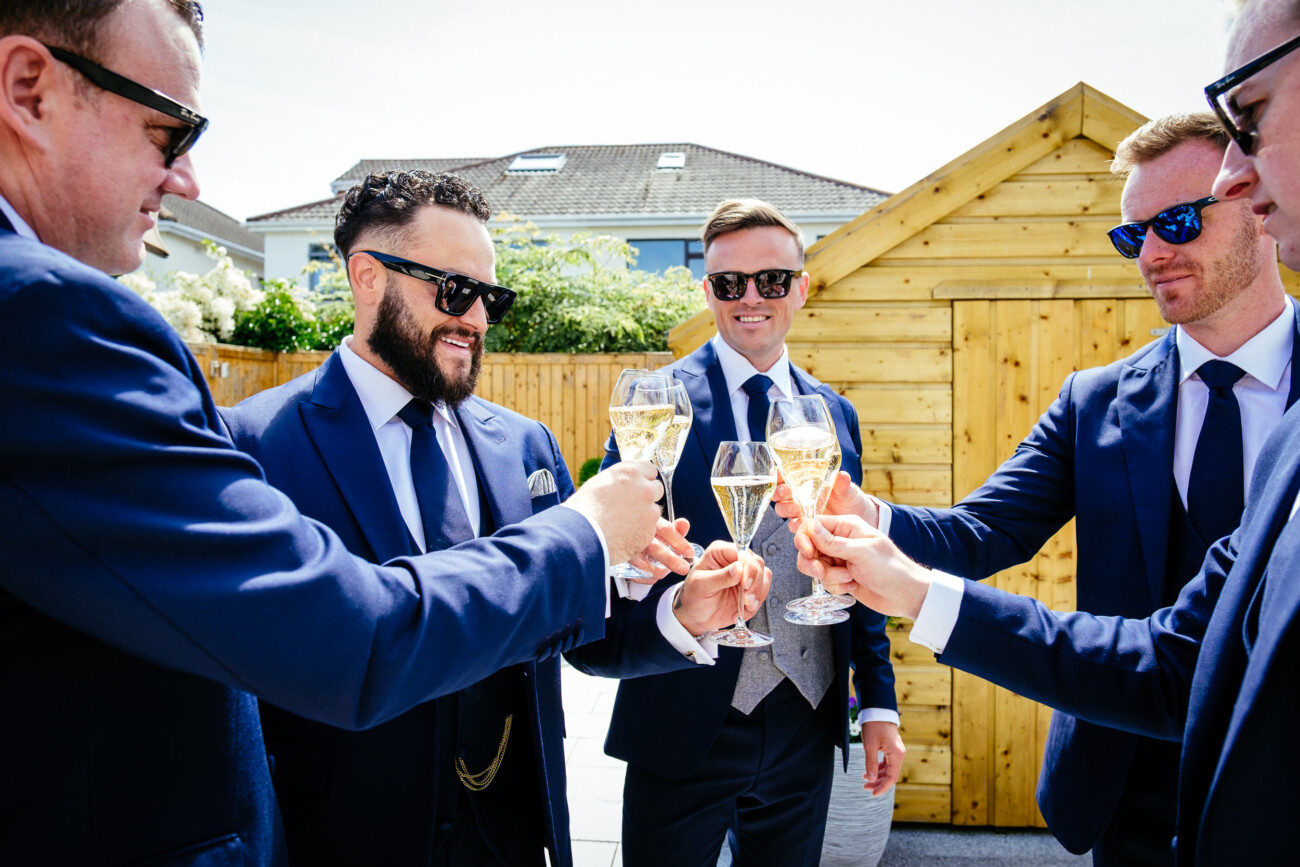 Tara and Darren’s Unforgettable Wedding at Bellingham Castle 27 Five men in suits and sunglasses toast with glasses of champagne outside, standing near a wooden shed on a sunny day during a wedding at Bellingham Castle.