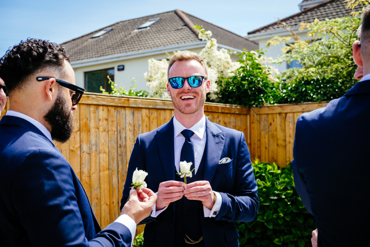 Tara and Darren’s Unforgettable Wedding at Bellingham Castle 28 Three men in suits stand outside by a wooden fence on a sunny day at a wedding at Bellingham Castle. The man in the centre smiles and holds a white buttonhole, while the other two men also wear buttonholes.