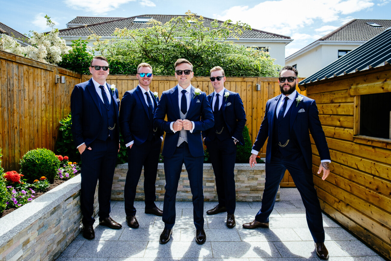 Tara and Darren’s Unforgettable Wedding at Bellingham Castle 32 Five men in dark suits and sunglasses pose together outdoors in a garden area with wooden fencing and a shed under a sunny sky, capturing a stylish moment during a wedding at Bellingham Castle.
