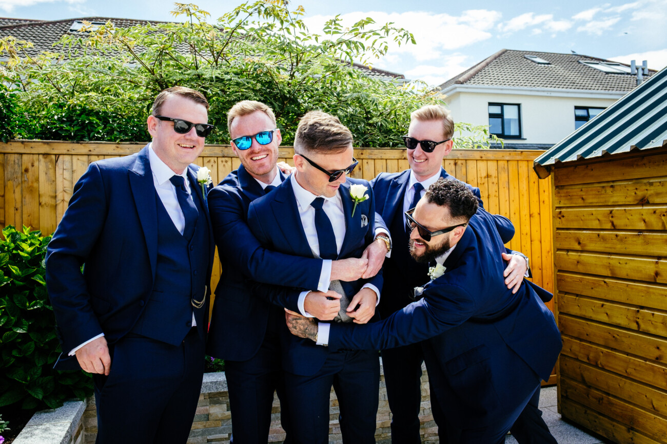 Tara and Darren’s Unforgettable Wedding at Bellingham Castle 33 Five men in blue suits and sunglasses stand outdoors, laughing and playfully hugging one man in the centre on a sunny day, with a wooden fence and greenery behind them—capturing a joyful moment during a wedding at Bellingham Castle.