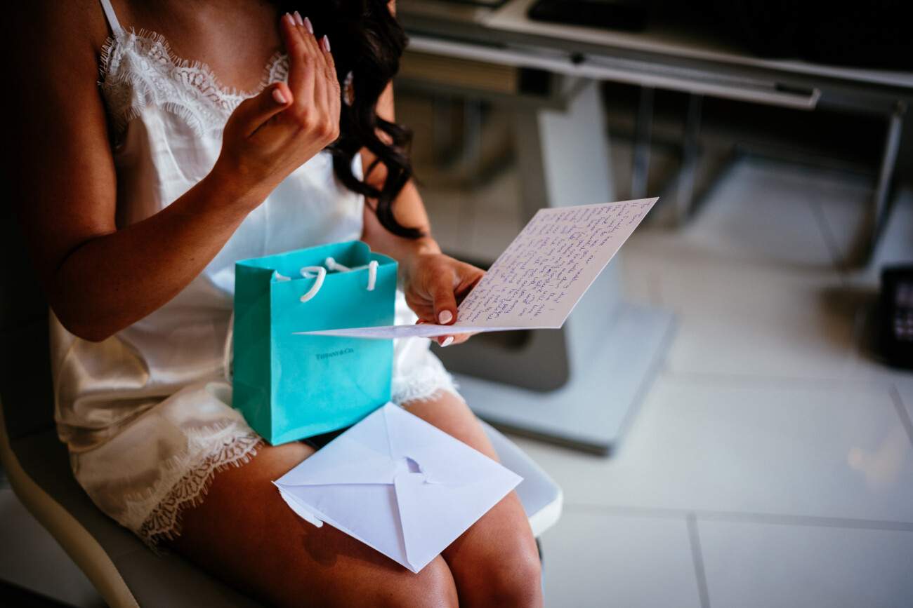 Tara and Darren’s Unforgettable Wedding at Bellingham Castle 34 A woman in a white petticoat sits holding a handwritten letter, with a turquoise gift bag and a white envelope on her lap, preparing for the wedding at Bellingham Castle.