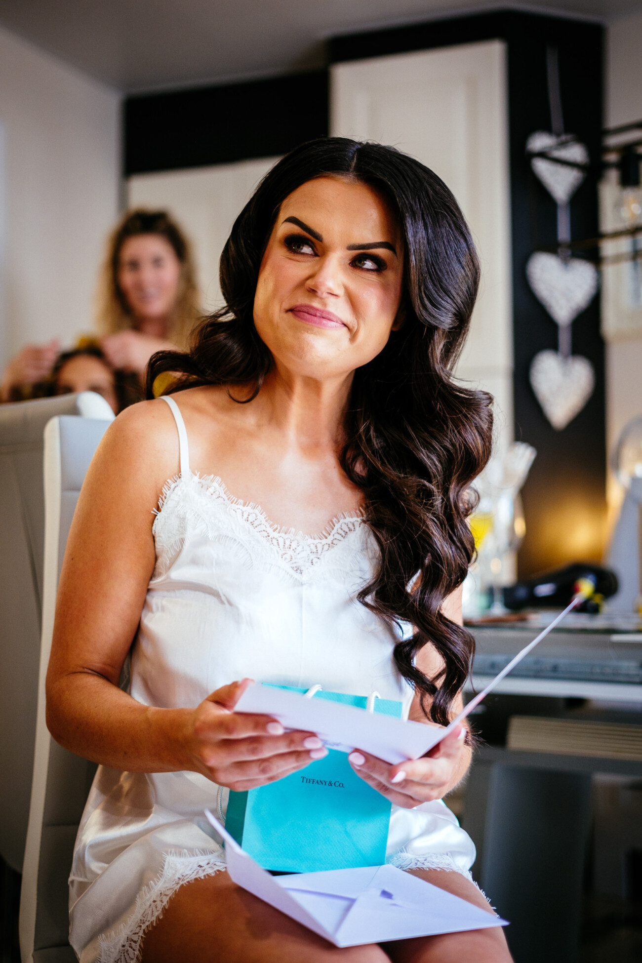 Tara and Darren’s Unforgettable Wedding at Bellingham Castle 35 Woman in a white slip dress sits indoors, smiling whilst holding an open Tiffany & Co. box and a card; the joyful atmosphere suggests preparations for a wedding at Bellingham Castle, as another person is blurred in the background.