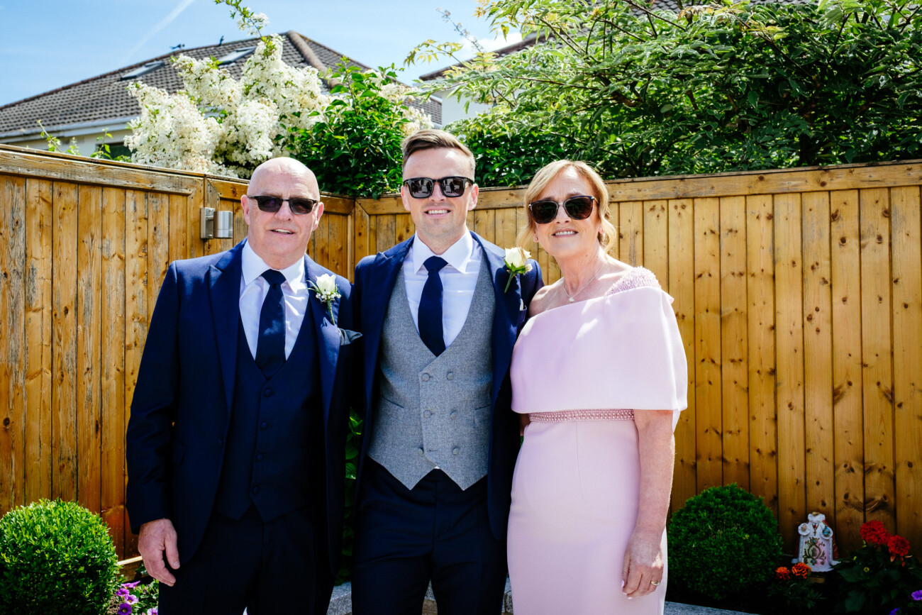 Tara and Darren’s Unforgettable Wedding at Bellingham Castle 36 Three adults dressed formally, standing together outdoors in front of a wooden fence and greenery at a wedding at Bellingham Castle, all wearing sunglasses and smiling at the camera.