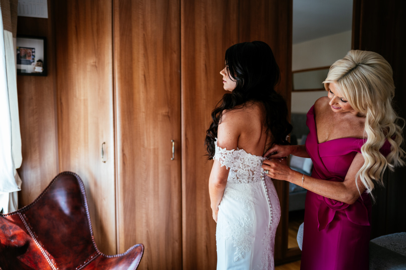 Tara and Darren’s Unforgettable Wedding at Bellingham Castle 39 A woman in a fuchsia dress helps another woman in a white lace wedding gown fasten the back of her dress in a wood-panelled room, capturing a tender moment before the wedding at Bellingham Castle.