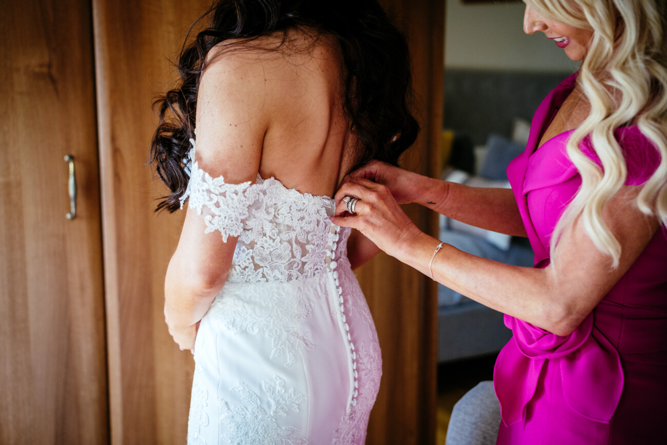 Tara and Darren’s Unforgettable Wedding at Bellingham Castle 40 A woman in a pink dress fastens the buttons on the back of another woman’s white lace wedding dress, preparing her for a beautiful wedding at Bellingham Castle.