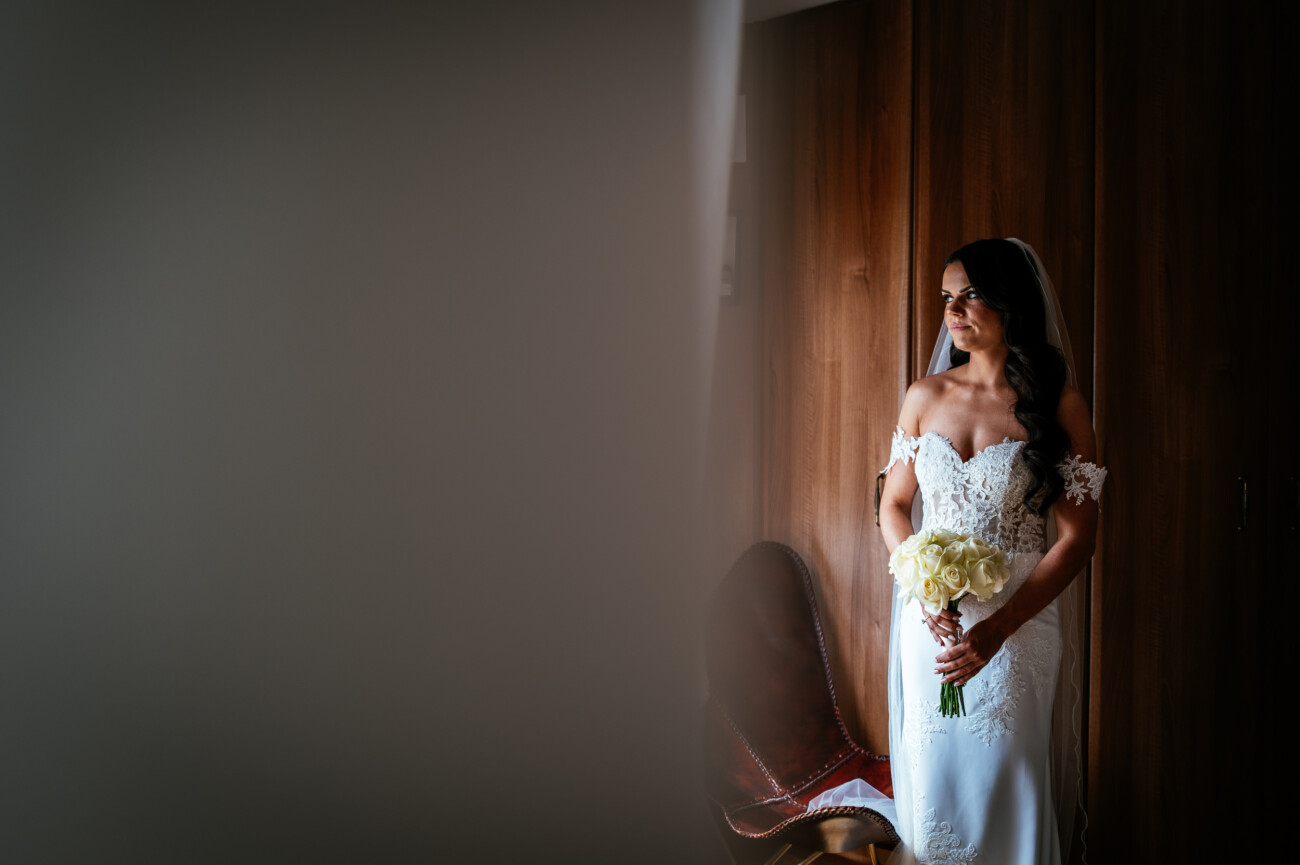 Tara and Darren’s Unforgettable Wedding at Bellingham Castle 41 A bride in a white dress holding a bouquet of flowers stands indoors near a wooden wall, looking to the side, capturing an elegant moment from a wedding at Bellingham Castle.