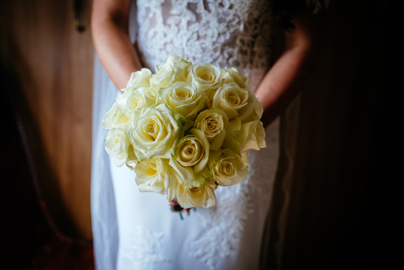Tara and Darren’s Unforgettable Wedding at Bellingham Castle 43 A person in a white lace dress holds a bouquet of cream-coloured roses, standing indoors with their hands clasped around the stems, ready to celebrate a wedding at Bellingham Castle.