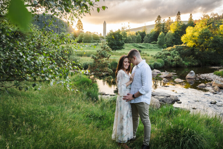 A couple stands closely together by a river in a green, grassy landscape at sunset, with trees and a tall stone tower in the background. Glendalough