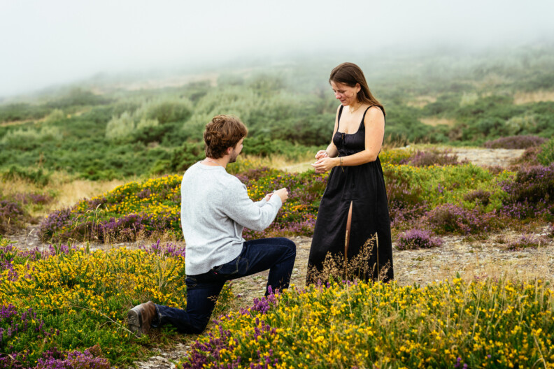 A man kneels on one knee, holding a ring while proposing to a woman in a black dress during an intimate Engagement Shoot in a field of wildflowers, with foggy hills setting the scene for Secret Proposals & Elopements in Ireland.