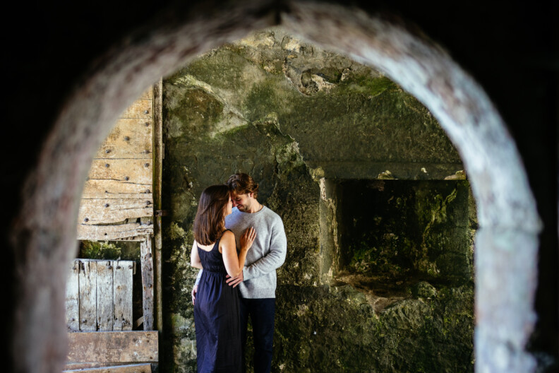 A man and woman stand closely together in a stone-walled room, framed by an arched doorway. Their contemplative pose captures the intimate essence of secret proposals & elopements in Ireland.