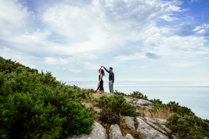 A couple stands on a rocky hilltop with greenery, holding hands as one twirls the other, under a partly cloudy sky with the ocean in the background—perfect for Secret Proposals & Elopements in Ireland.