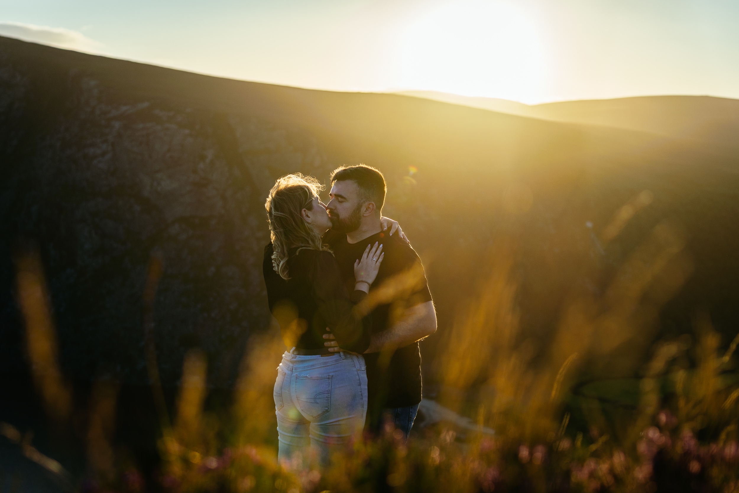 A couple embraces on a hillside at sunset, sunlight illuminating their faces and tall grass in the foreground—perfect for Engagement Shoots or Secret Proposals & Elopements in Ireland.