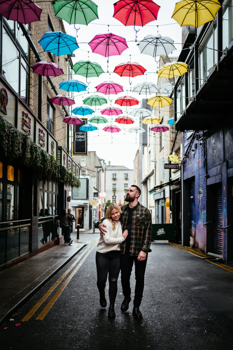 A couple walks arm in arm down a city street under colorful umbrellas suspended above the road on a cloudy day, capturing a moment perfect for Engagement Shoots or even Secret Proposals & Elopements in Ireland.