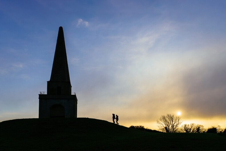 Silhouette of two people walking near an obelisk-shaped tower on a hill at sunset, with trees and a colorful sky in the background—an idyllic spot for Secret Proposals & Elopements in Ireland.