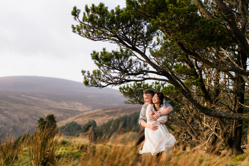 A man and woman embrace and smile under a tree on a grassy hillside with mountains in the background, capturing the magic of Engagement Shoots and Secret Proposals & Elopements in Ireland.