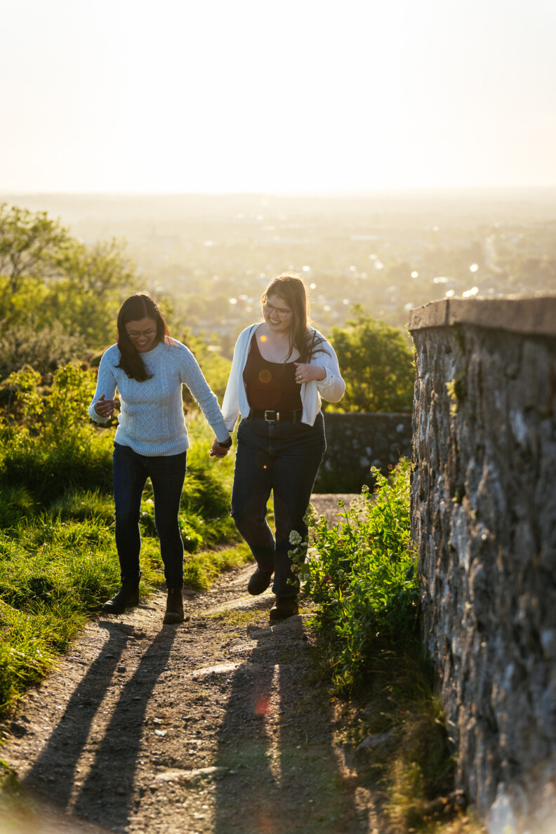 Two women walk hand in hand up a sunlit path beside a stone wall, surrounded by greenery and a bright sky—an idyllic setting for engagement shoots or secret proposals & elopements in Ireland.