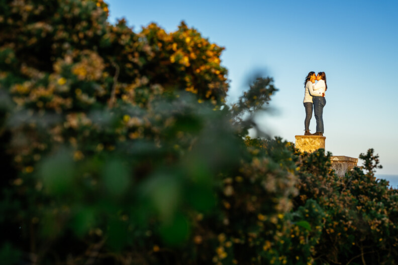 Two people stand close together on a stone platform, surrounded by green bushes and flowers under a clear blue sky—the perfect setting for Secret Proposals & Elopements in Ireland or romantic Engagement Shoots.