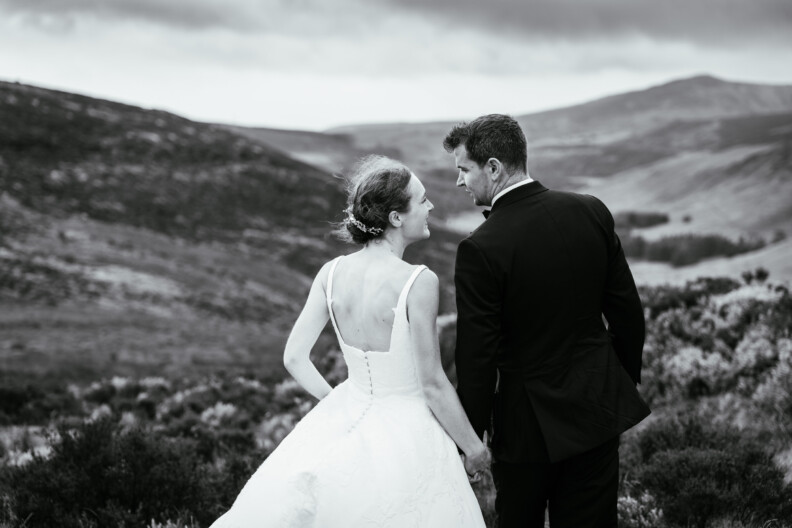 A bride and groom stand side by side outdoors, facing away from the camera, with rolling hills in the background under a cloudy sky—perfect inspiration for Secret Proposals & Elopements in Ireland.