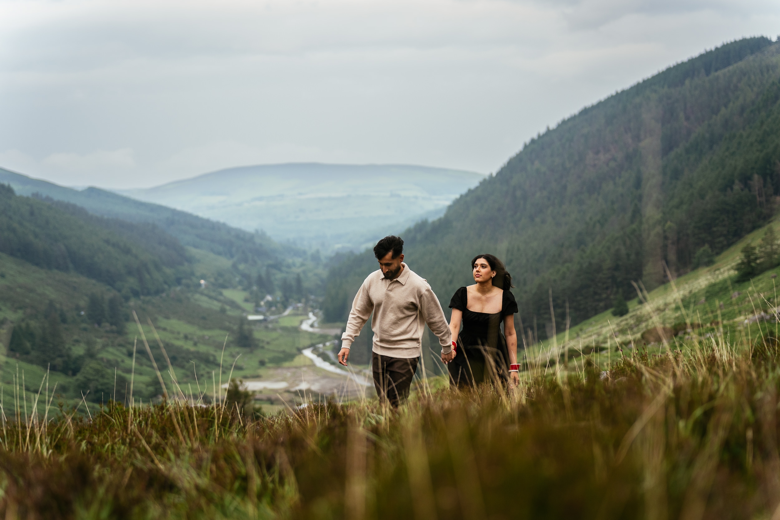 A couple walks hand in hand through a grassy hillside with mountains and a valley in the background under a cloudy sky—perfect for magical Engagement Shoots or intimate Secret Proposals & Elopements in Ireland.