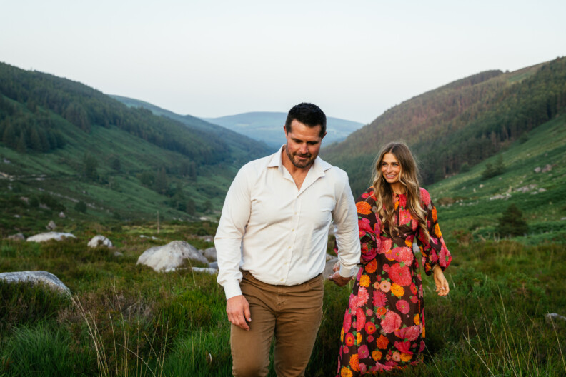A man in a white shirt and brown pants holds hands with a smiling woman in a floral dress as they walk through a green valley, perfect for Engagement Shoots or Secret Proposals & Elopements in Ireland, with hills and large rocks behind them.