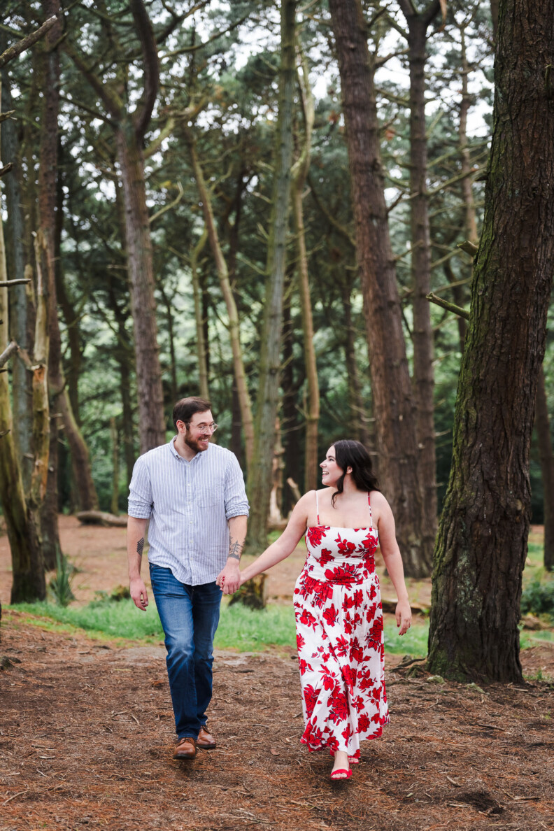 A man and woman walk hand-in-hand through a forest during their Engagement Shoot. The man wears jeans and a striped shirt; the woman wears a white dress with red floral patterns. Trees surround them, creating a romantic atmosphere.