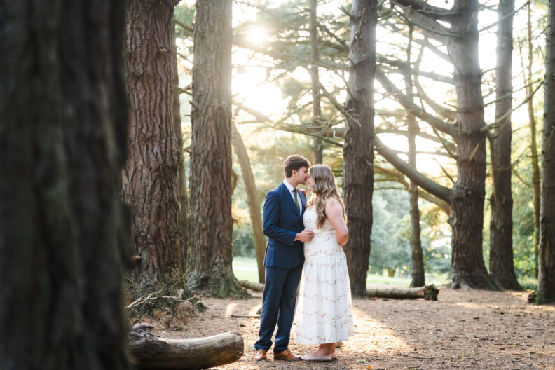 A man in a suit and a woman in a white dress stand close together in a sunlit forest, surrounded by tall trees—perfect for engagement shoots or secret proposals & elopements in Ireland.
