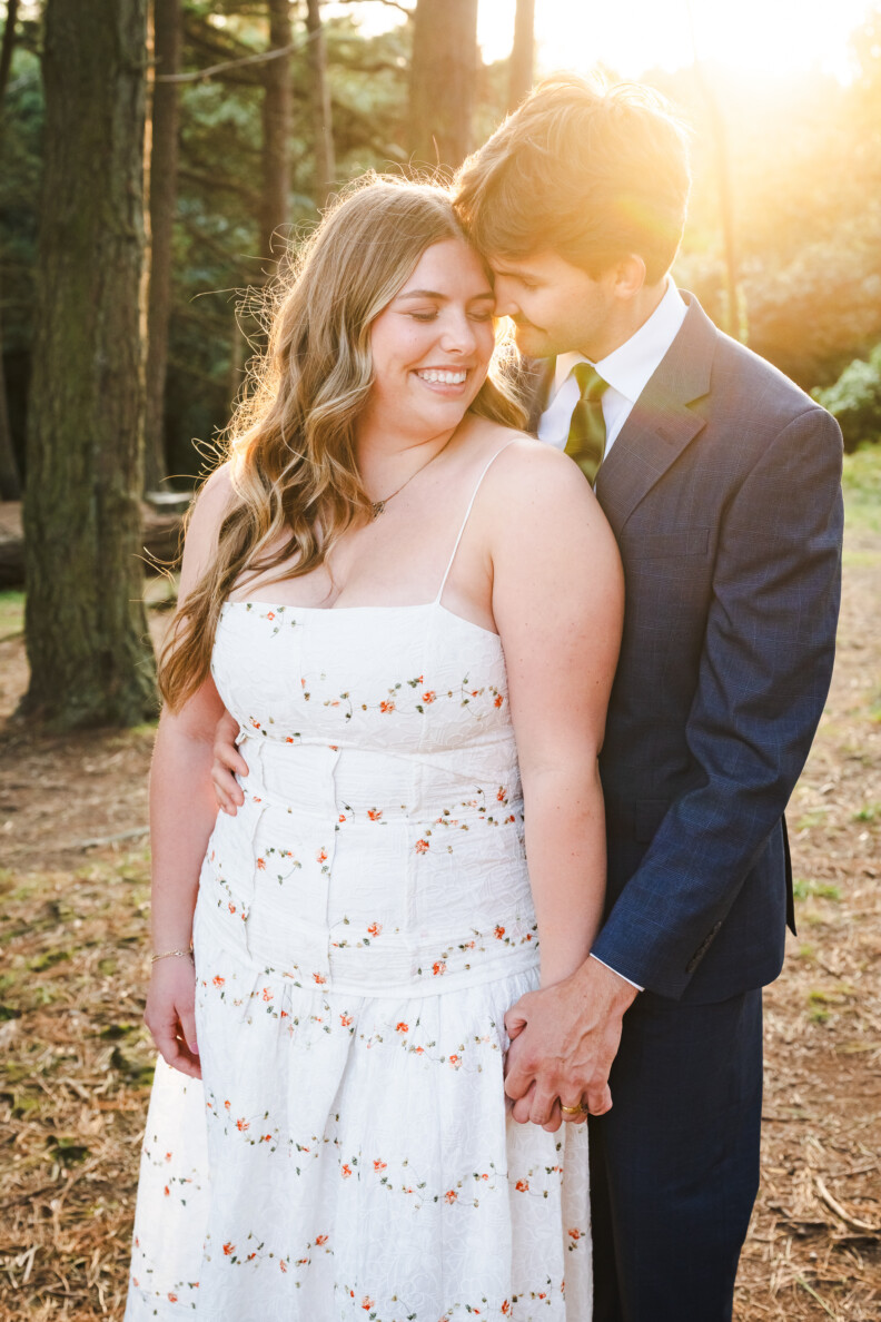 A man in a suit stands behind a woman in a white dress, holding her hand and kissing her temple in a sunlit wooded area—capturing the romance of Secret Proposals & Elopements in Ireland.
