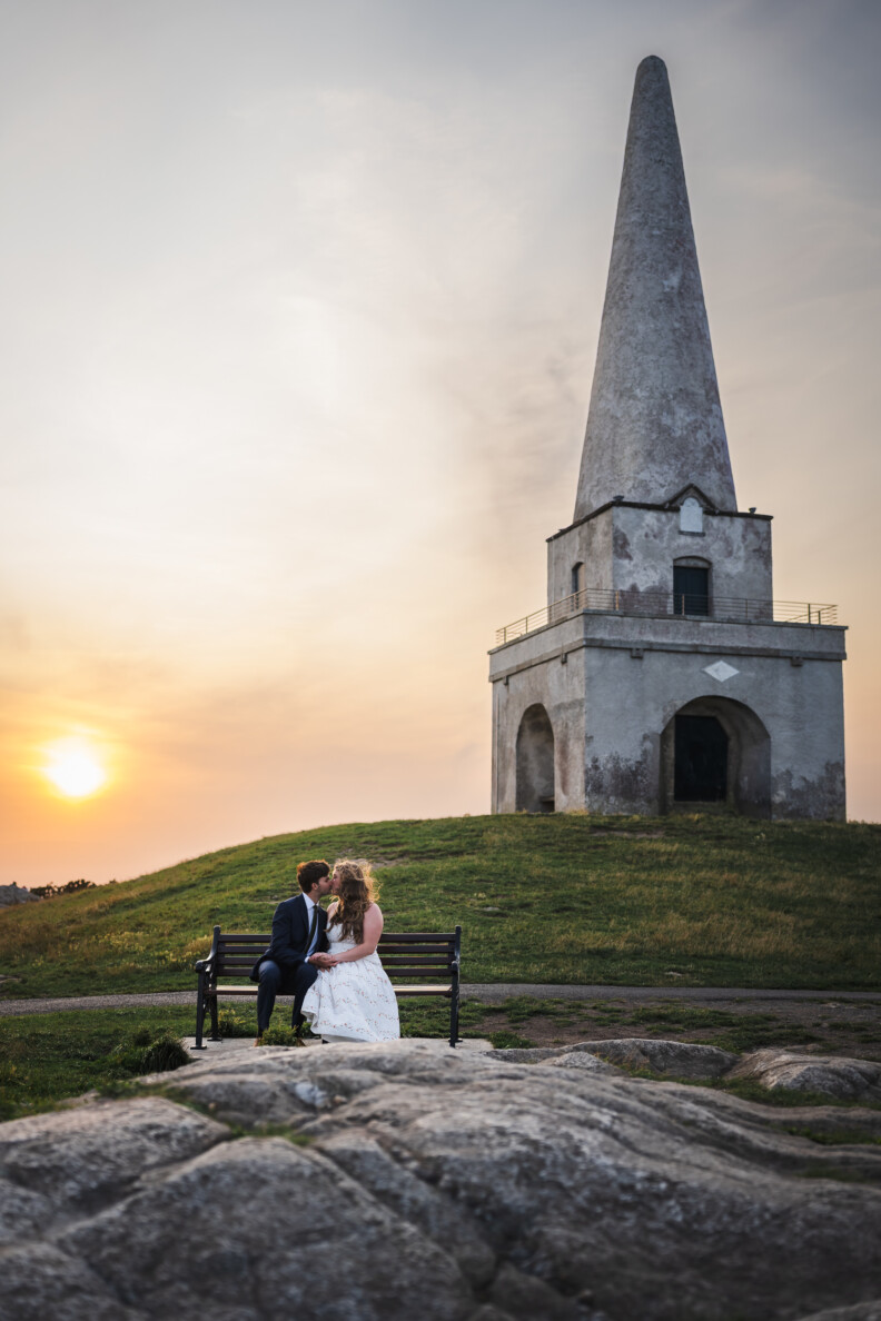 A couple in formal attire sits on a bench at sunset near a historic stone tower with a tall spire on a grassy hill, perfect for magical Engagement Shoots and Secret Proposals & Elopements in Ireland.