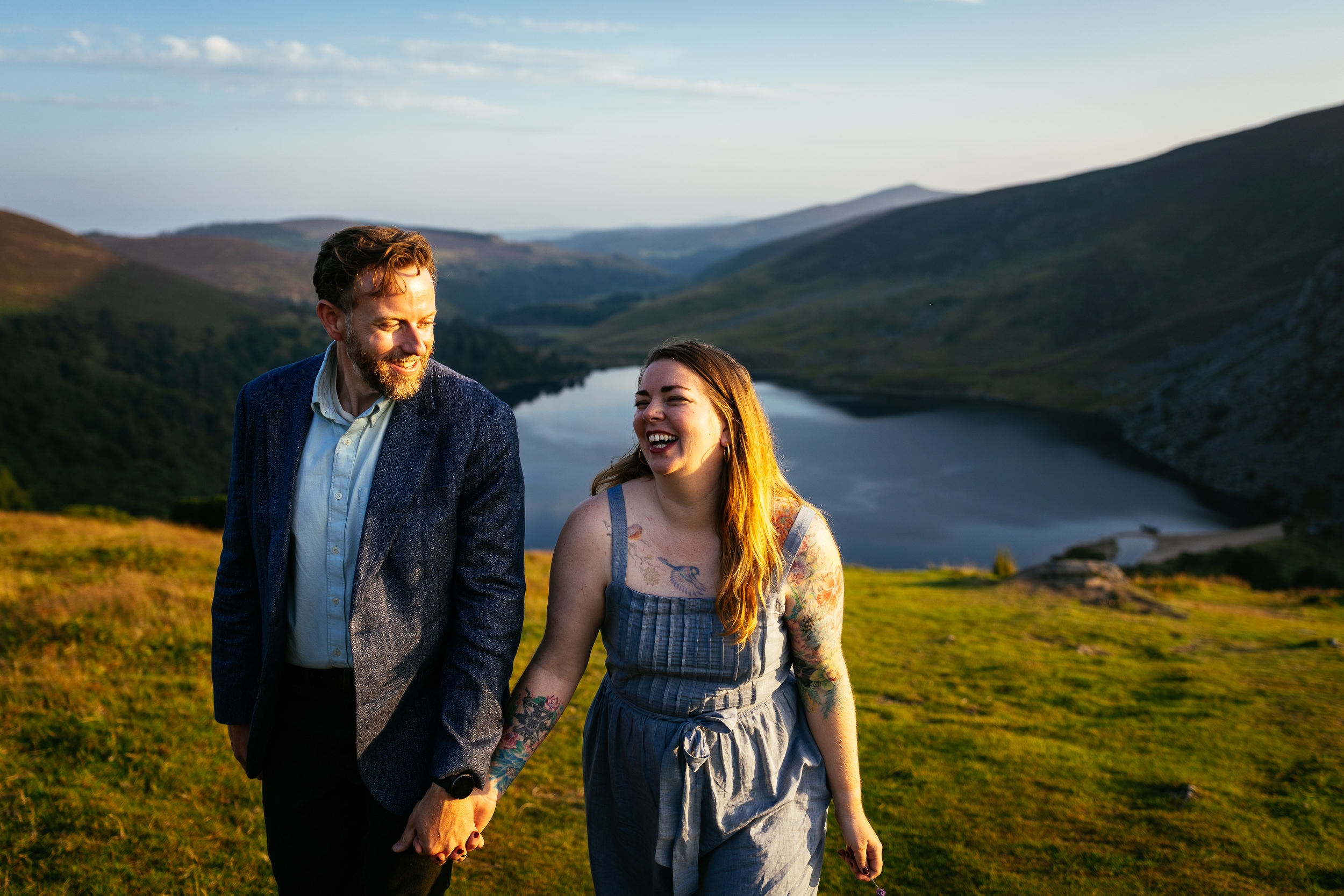 A man and a woman holding hands and smiling while walking on grass with a lake and hills in the background—perfect for capturing Engagement Shoots or Secret Proposals & Elopements in Ireland.