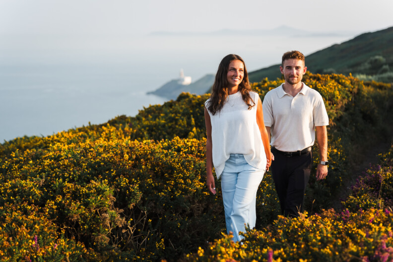 A woman and a man walk along a path surrounded by green bushes and yellow flowers, with the sea and distant hills in the background—an idyllic setting for Engagement Shoots or Secret Proposals & Elopements in Ireland.
