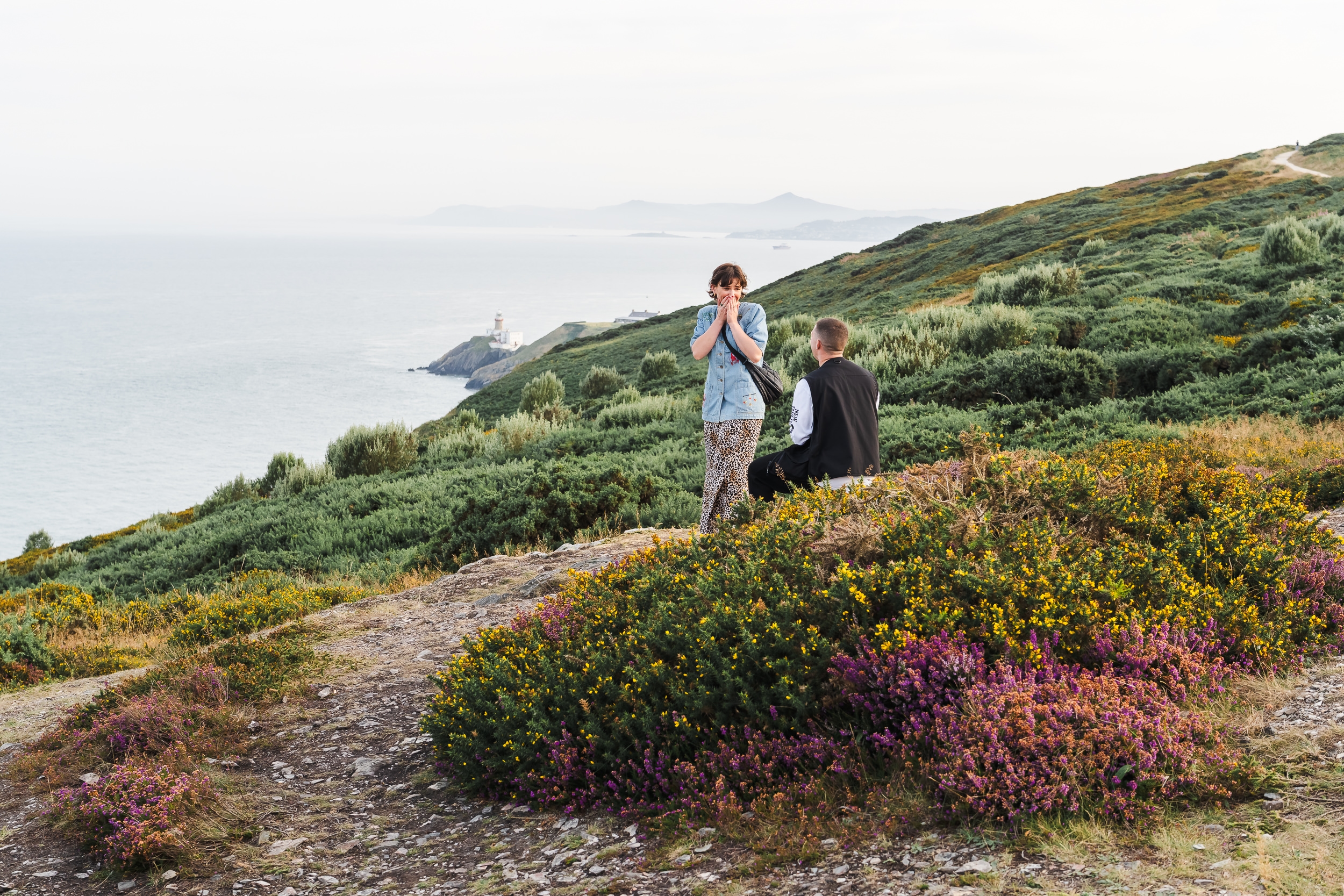Two people are on a hillside covered in green and flowering shrubs, with one standing and one sitting, overlooking the sea and a distant lighthouse—an idyllic spot for secret proposals & elopements in Ireland.