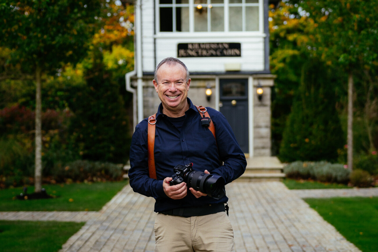 David Duignan photographer stands on a stone pathway holding a camera, embodying the spirit of a documentary wedding photographer in Ireland, with a building and lush greenery in the background.